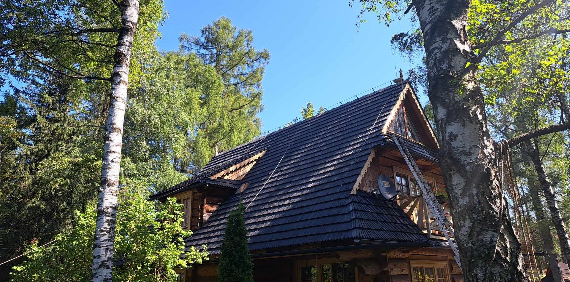 Dom w stylu góralskim z nowym, ciemnym dachem z drewnianych gontów, z drabiny opartą o balkon na poddaszu, otoczony drzewami, w słoneczny dzień.