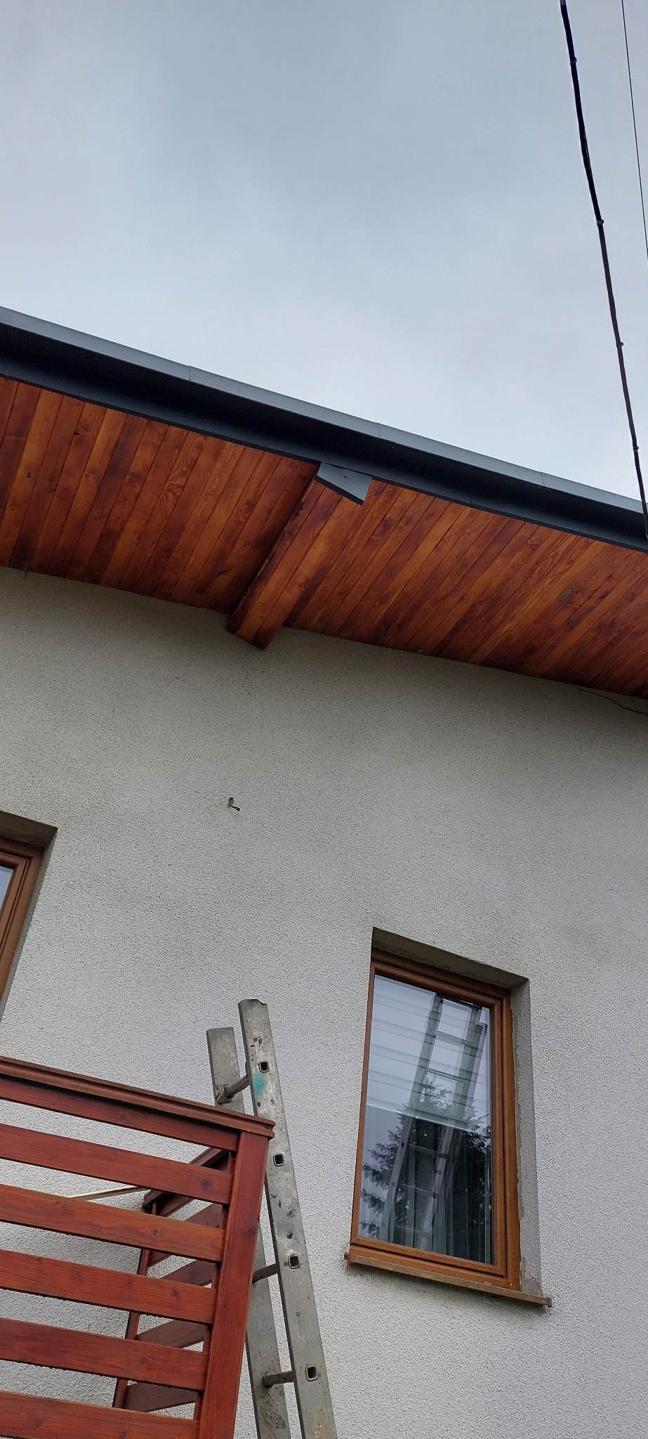 Drewniana podbitka dachowa w kolorze orzecha, zamontowana na elewacji domu z oknami, widoczna drabina oparta o ścianę.