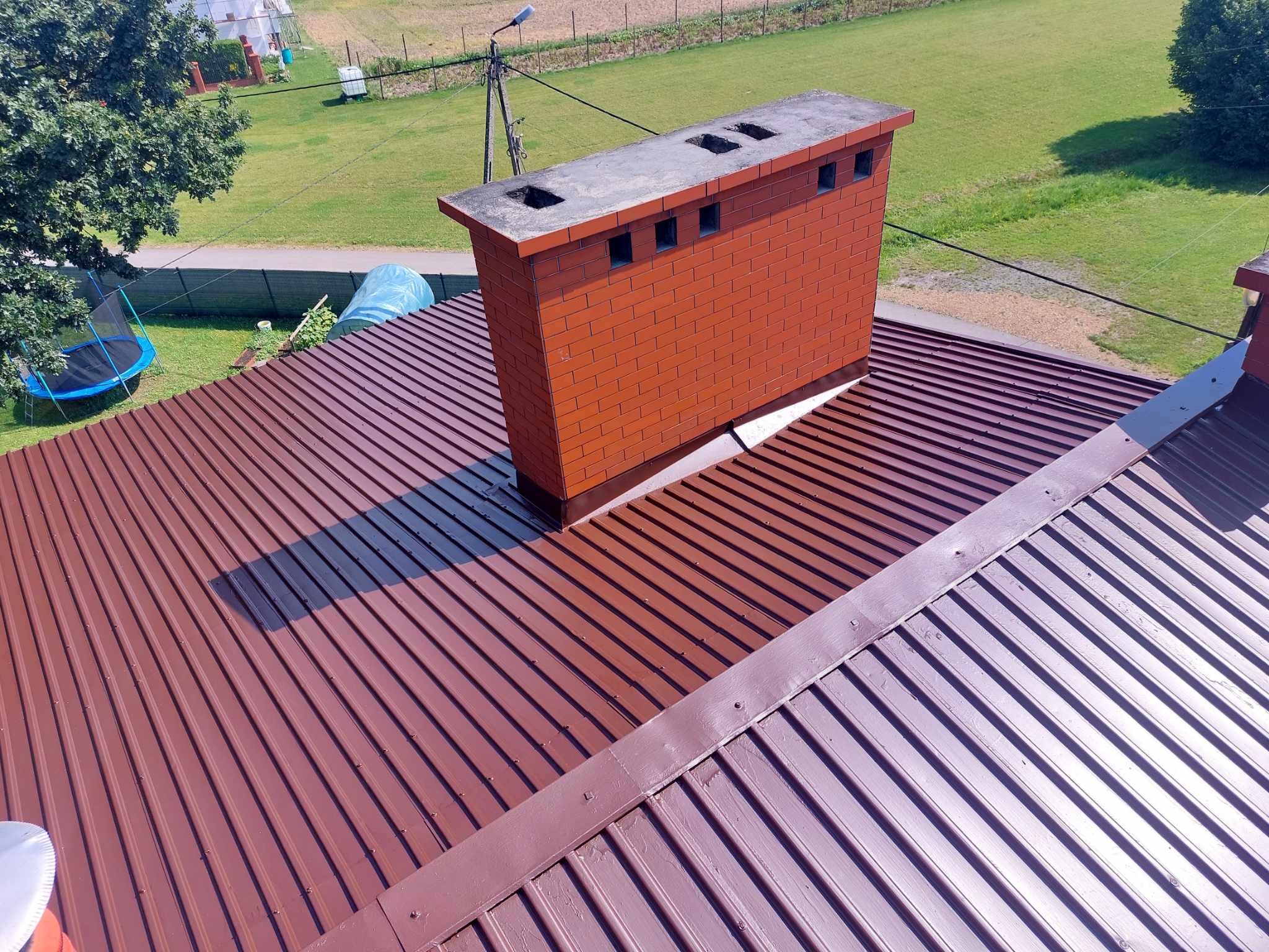 Czerwony dach z blachy trapezowej po renowacji, widoczny ceglany komin z betonową czapą, cień komina na dachu, w tle trawnik i zabudowania.