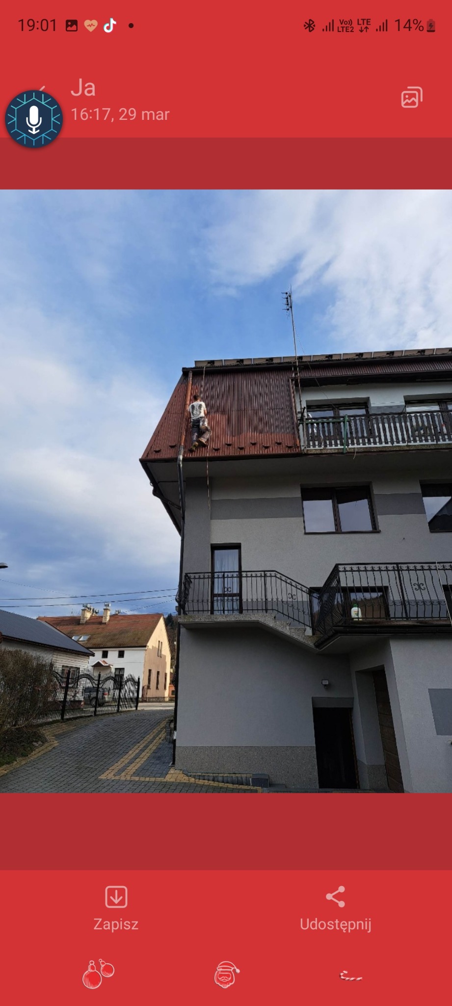 Pracownik na dachu z blachy trapezowej, montaż anteny na tle błękitnego nieba i zabudowy jednorodzinnej.