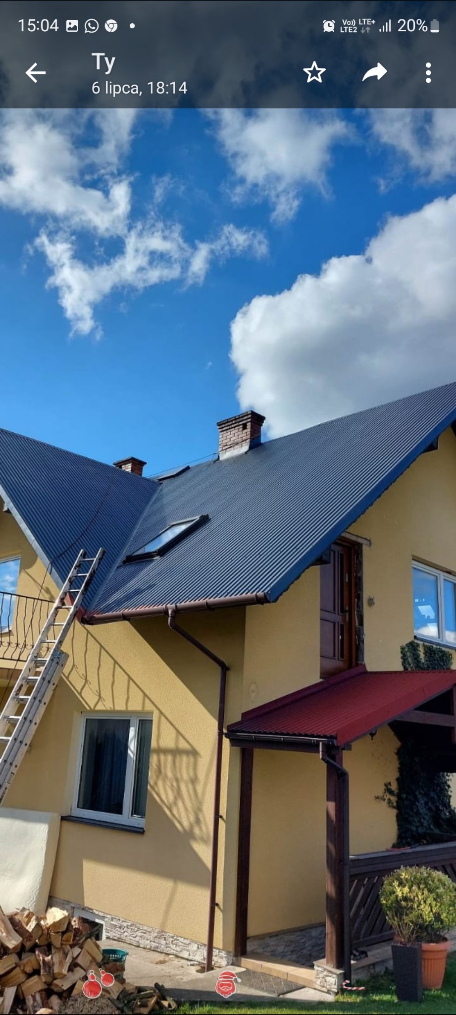 Dom z ciemnym dachem z blachy trapezowej, widoczne okna dachowe, komin z cegły, elewacja w kolorze piaskowym, drabina oparta o rynnę, składowane drewno opałowe.