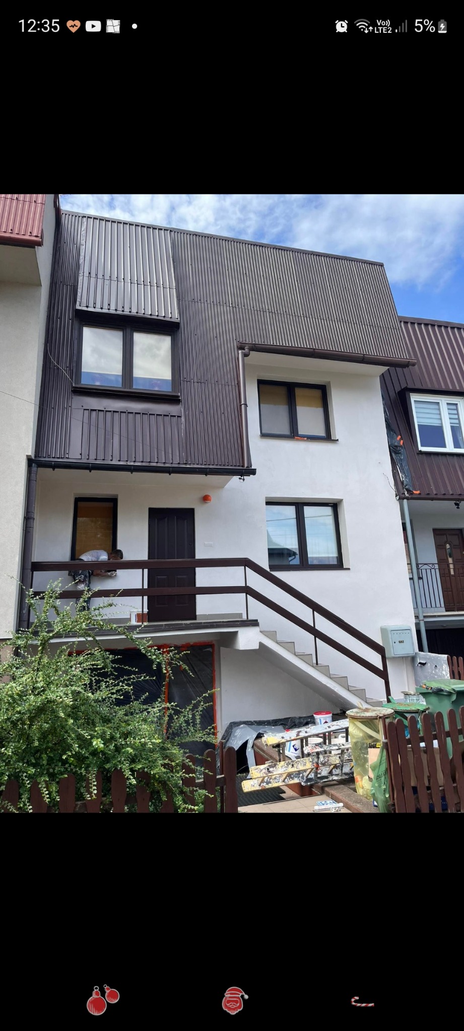 Dwupiętrowy dom z białymi ścianami i brązowym dachem z blachy trapezowej, widoczne prace na balkonie i drabina oparta o ogrodzenie, sugerujące remont elewacji.