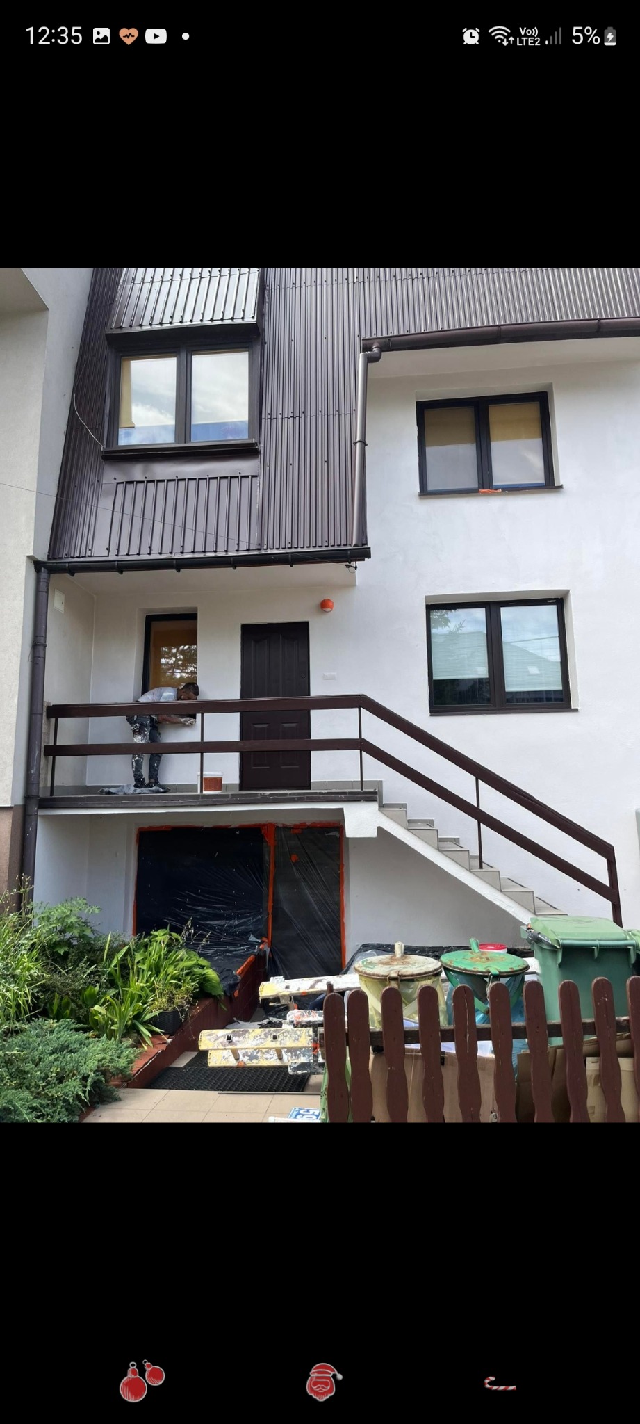 Pracownik maluje białą elewację domu z brązowymi oknami i dachem, zabezpieczone folią drzwi wejściowe na parterze.