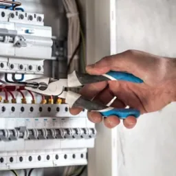 Elektryk przy pracy: ręka z kombinerkami tnie przewód w skrzynce rozdzielczej z bezpiecznikami, widoczne czerwone i czarne kable.