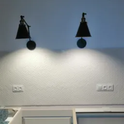 Biała mozaika ułożona w jodełkę na ścianie nad blatem kuchennym, z dwoma czarnymi lampami ściennymi i gniazdkami elektrycznymi, widoczne okno i fragment mebli kuchennych w trakcie montażu.