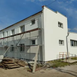 Dwupiętrowy budynek z białą elewacją, częściowo pokryty rusztowaniami, z widocznymi materiałami budowlanymi i drewnianymi deskami na ziemi, pod błękitnym niebem.