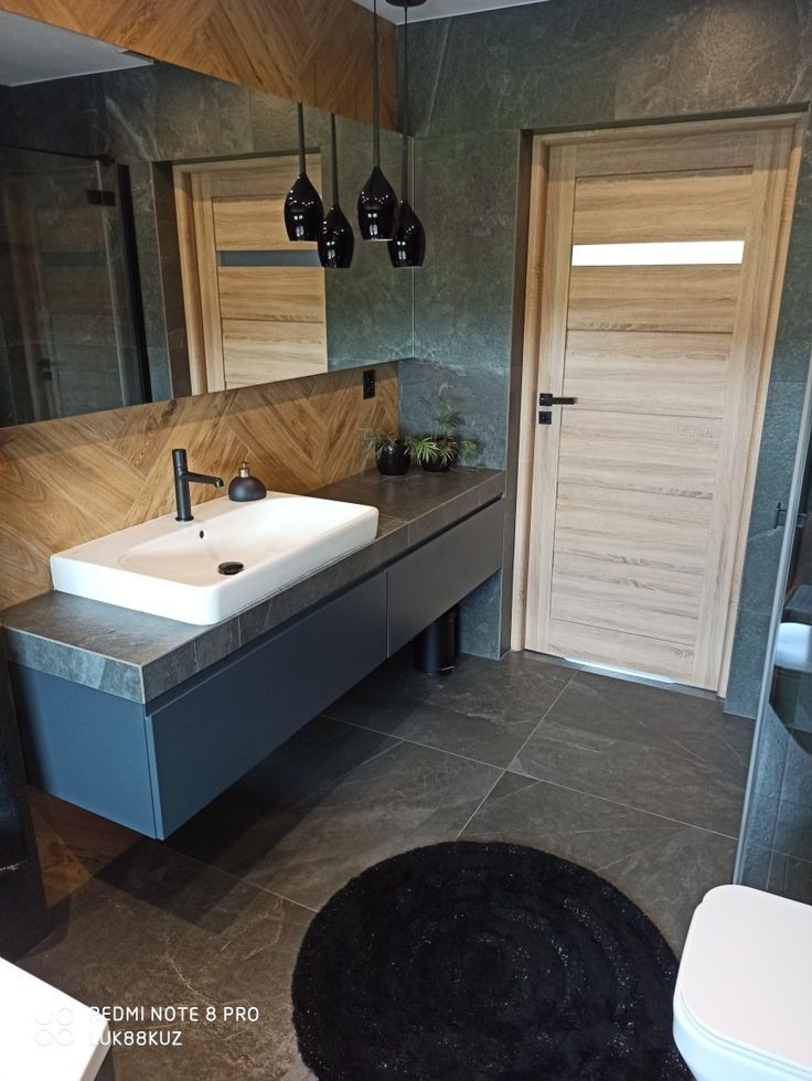 Nowoczesna łazienka z ciemnoszarą płytką, drewnianymi akcentami, wiszącą szafką z umywalką nablatową, czarną armaturą i okrągłym, czarnym dywanikiem.