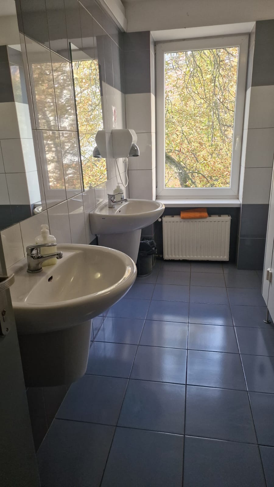 Umywalki w toalecie publicznej z dozownikami mydła i ręczników, okno z widokiem na drzewo, pomarańczowy ręcznik na grzejniku.