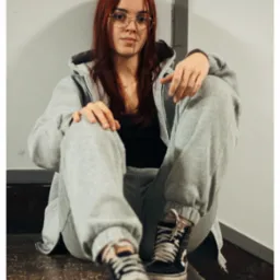 Portret młodej kobiety w okularach, siedzącej na schodach w szarym dresie i czarnych trampkach, z rudymi włosami opadającymi na ramiona.