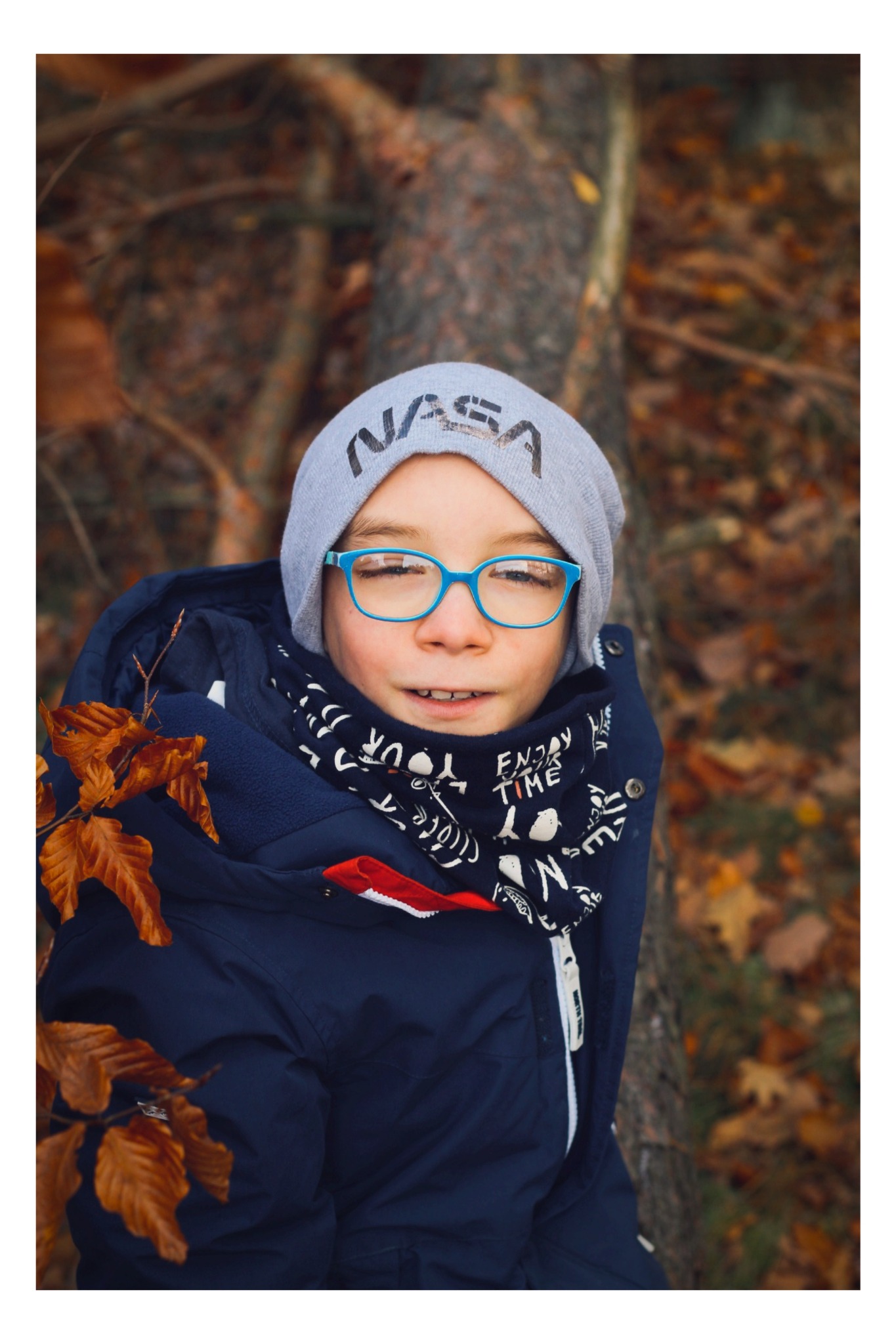 Portret uśmiechniętego chłopca w niebieskich okularach i czapce z napisem NASA, ubrany w granatową kurtkę i szalik w jesiennym plenerze.