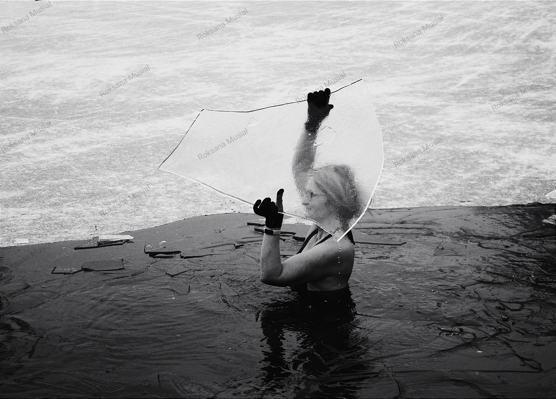 Czarno-białe zdjęcie kobiety zanurzonej w wodzie, trzymającej w dłoniach fragment lodu na tle zamarzniętego jeziora. Widoczne odbicie sylwetki w wodzie.