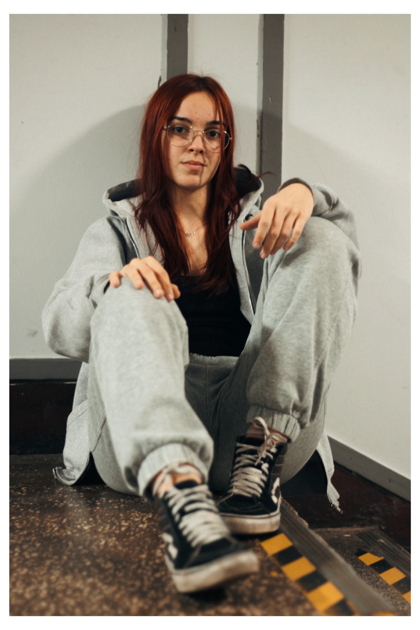 Portret młodej kobiety w okularach, siedzącej na schodach w szarym dresie i czarnych trampkach, z rudymi włosami opadającymi na ramiona.