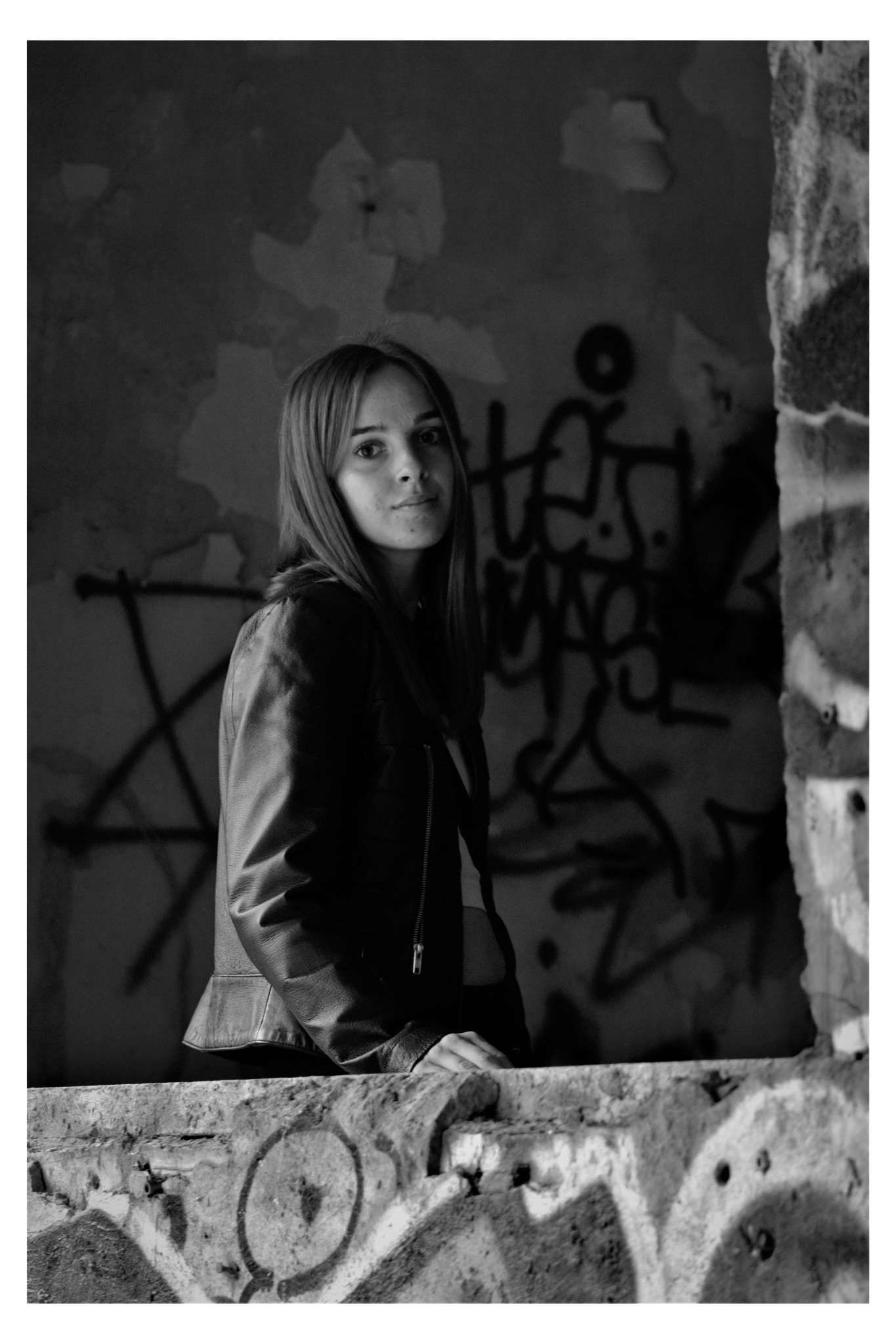Portret młodej kobiety w skórzanej kurtce, opierającej się o zniszczony, betonowy mur z graffiti, w czarno-białej tonacji.