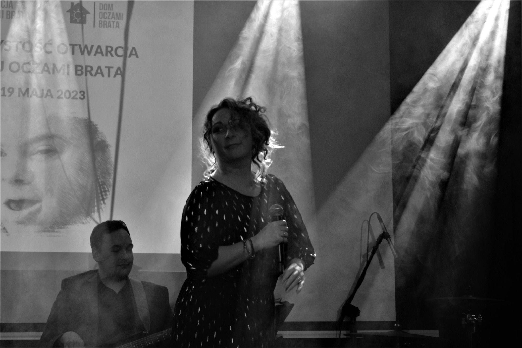 Czarno-białe zdjęcie kobiety z mikrofonem na scenie, oświetlonej reflektorami, w tle widoczny muzyk z gitarą i plakat z napisem 'Dom Oczami Brata' i datą 19 maja 2023.