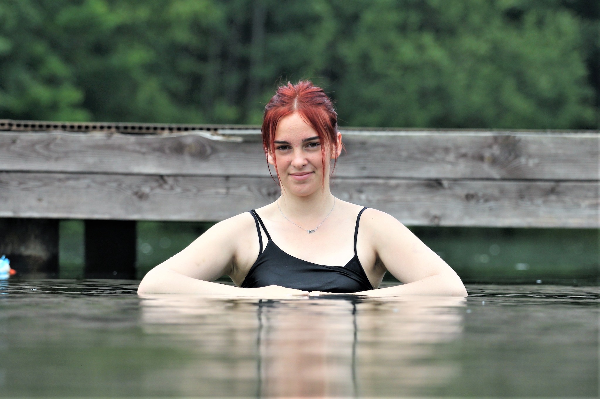 Portret uśmiechniętej kobiety w czarnym stroju kąpielowym, zanurzonej w wodzie po ramiona, z rękami opartymi o jej powierzchnię, na tle drewnianego pomostu i zieleni drzew.