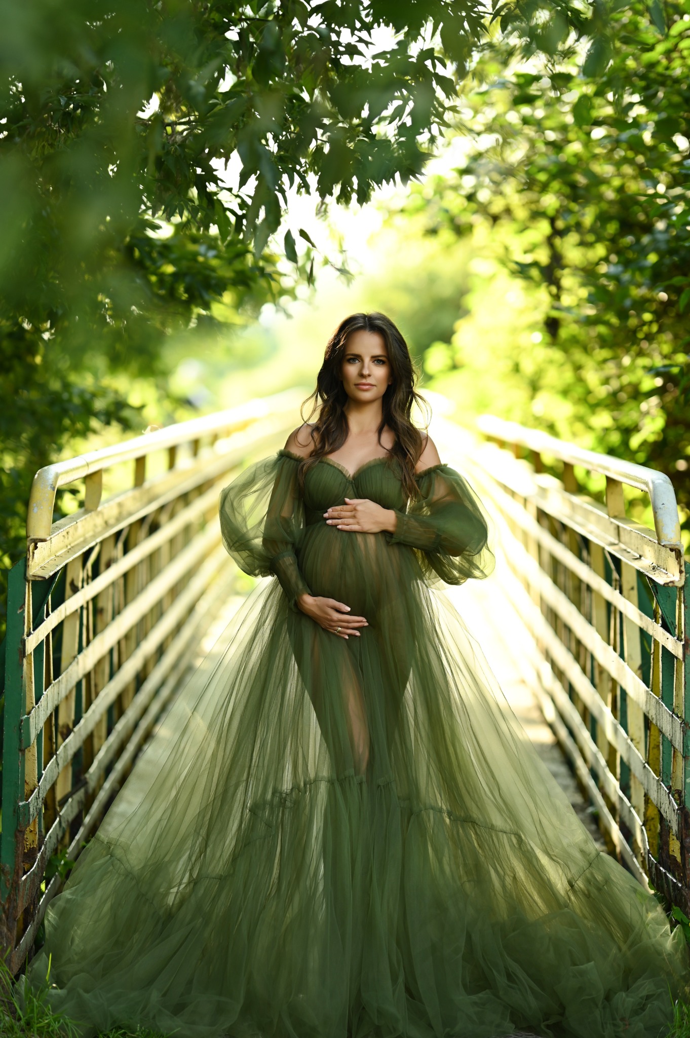 Kobieta w ciąży w zwiewnej, zielonej sukni pozuje na starym moście w Lublinie, otoczona zielenią drzew.
