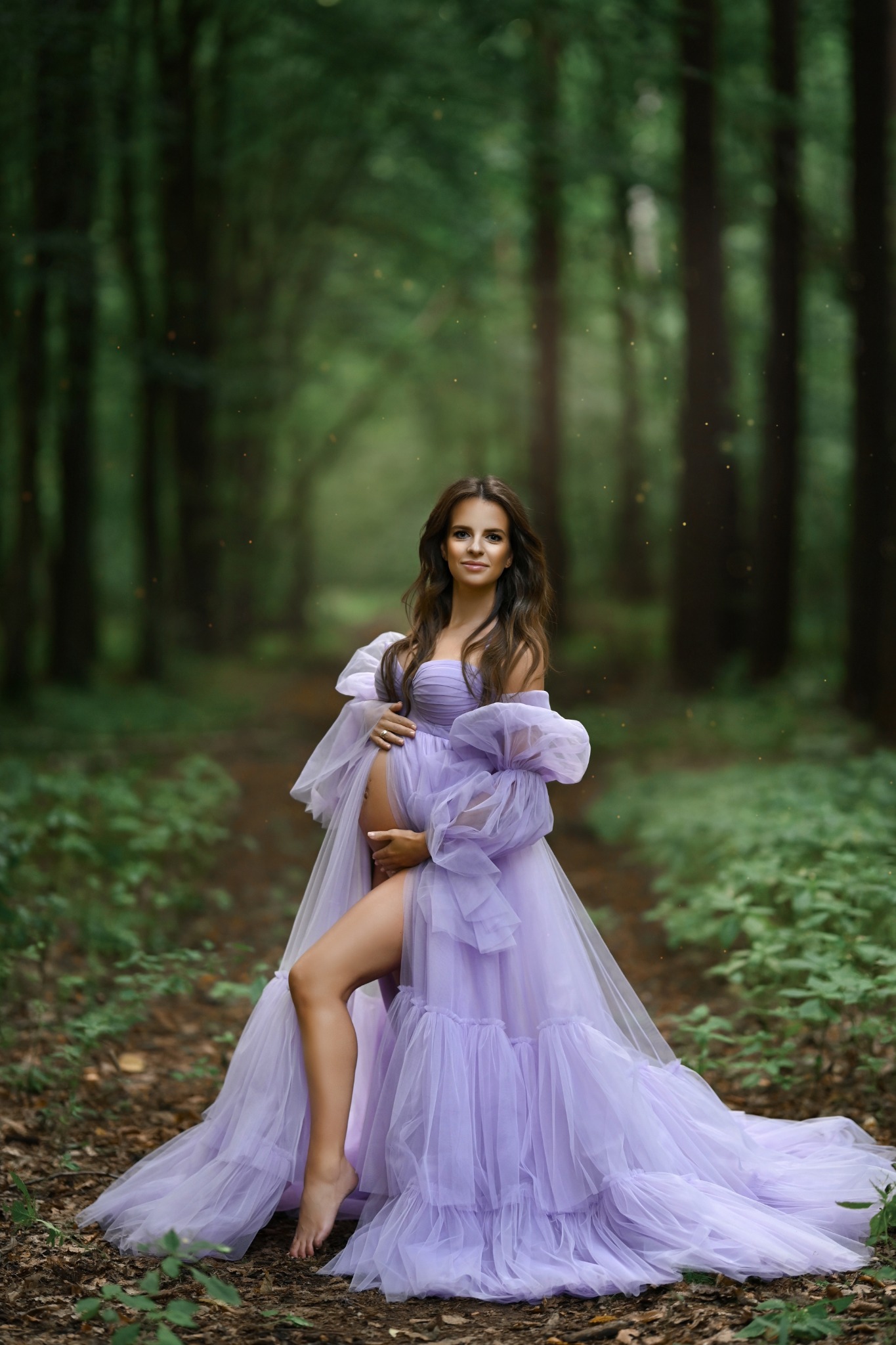 Kobieta w zwiewnej, fioletowej sukni ciążowej pozuje w lesie z delikatnym światłem, eksponując brzuszek i bose stopy na leśnym podłożu.