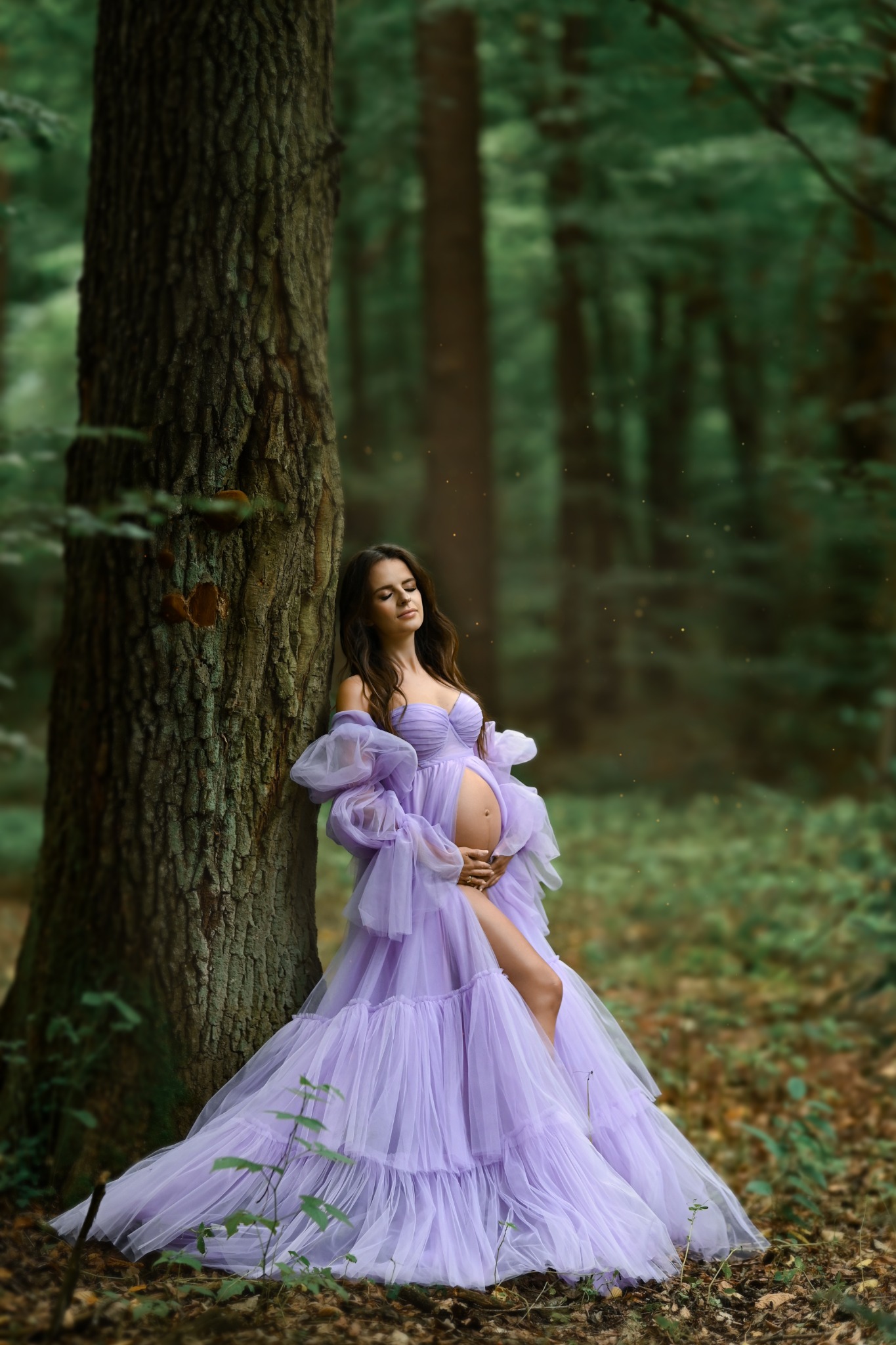 Kobieta w zwiewnej, liliowej sukni ciążowej opiera się o drzewo w lesie, z zamkniętymi oczami i dłońmi na brzuchu.