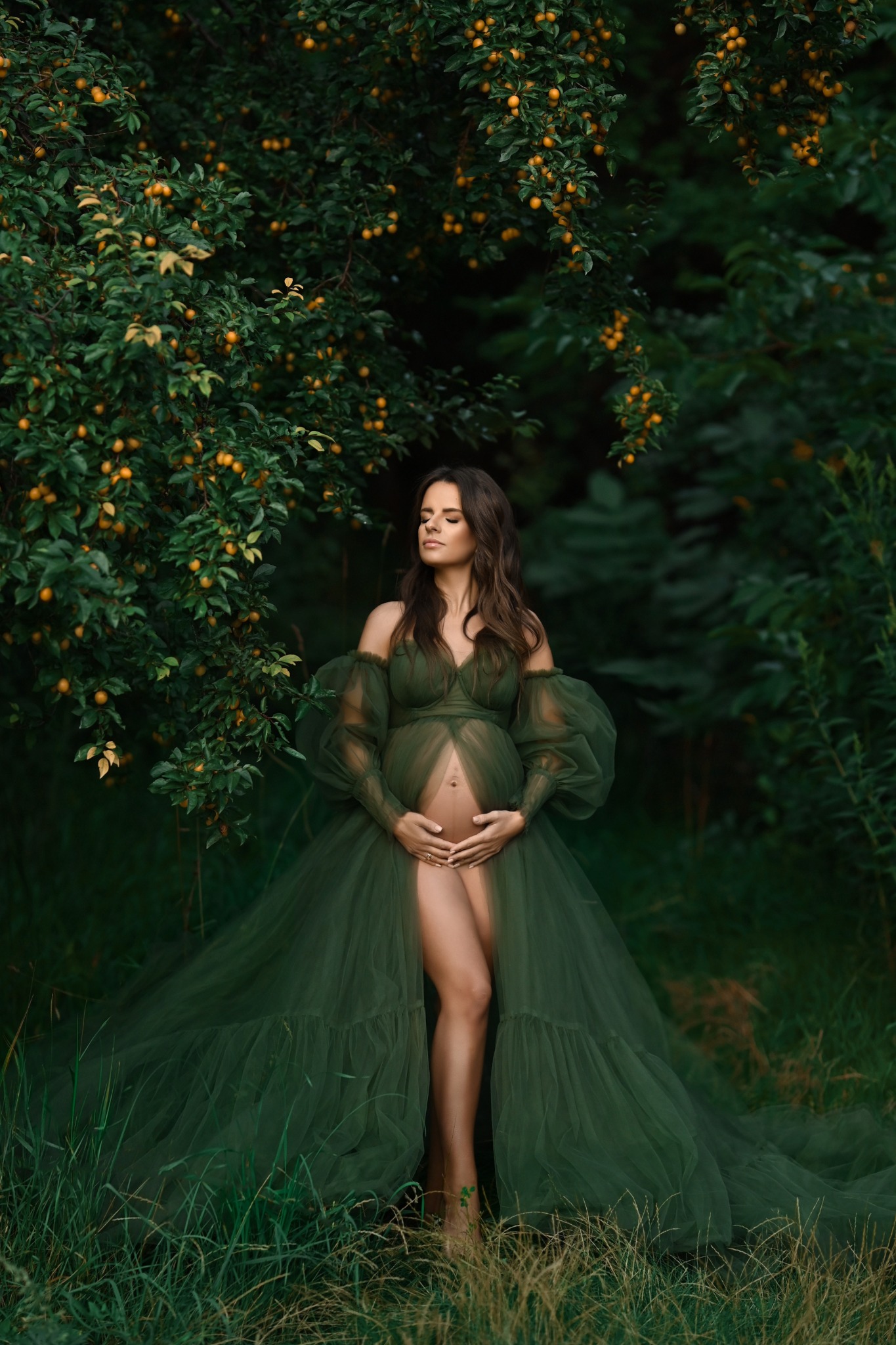 Kobieta w ciąży w długiej, zielonej sukni pozuje na łonie natury pod drzewem z żółtymi owocami, trzymając ręce na brzuchu i mając zamknięte oczy.