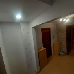 Przedpokój po odświeżeniu, ściany pomalowane na biało z rzędem okrągłych włączników, drewniane szafki na buty, drzwi wejściowe w kolorze ciemnego brązu, fragment ściany wykończony dekoracyjnym...