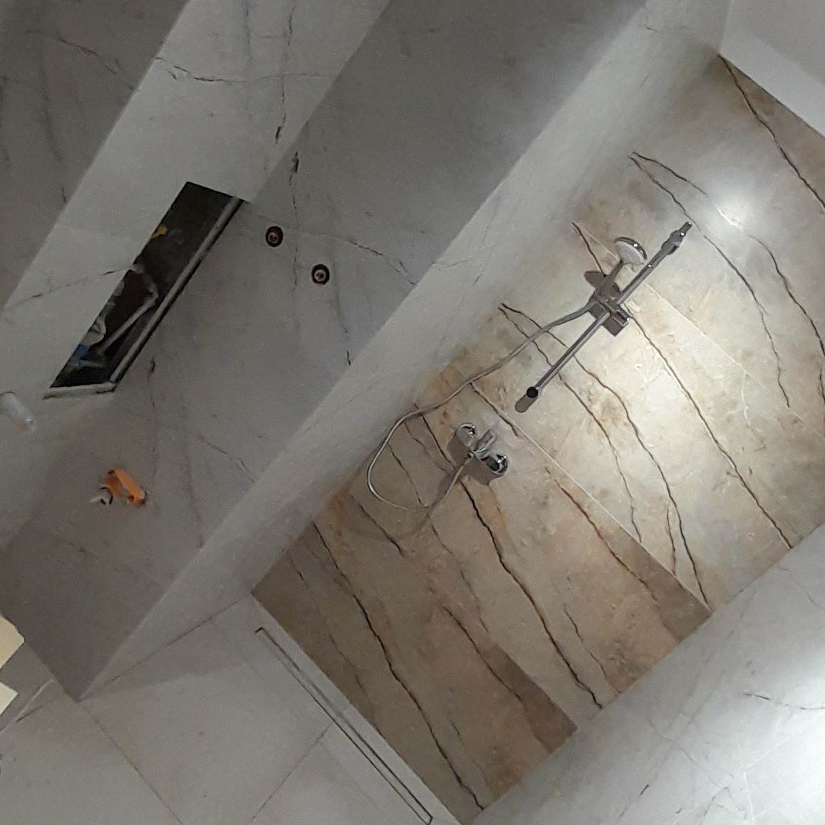 Wnętrze łazienki w trakcie prac wykończeniowych: zamontowana bateria prysznicowa, widoczny otwór w suficie z wystającymi przewodami, marmurowe płytki ścienne w odcieniach beżu i szarości.