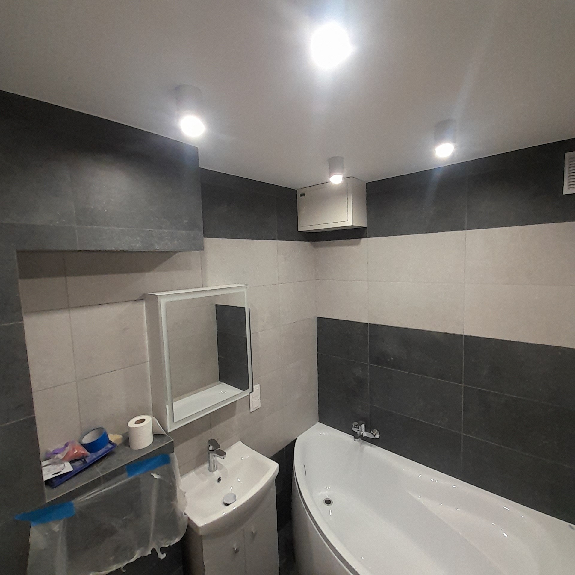 Wnętrze łazienki w trakcie remontu, widoczne płytki w odcieniach szarości i bieli, wanna, umywalka z szafką, lustro oraz akcesoria malarskie zabezpieczone folią.