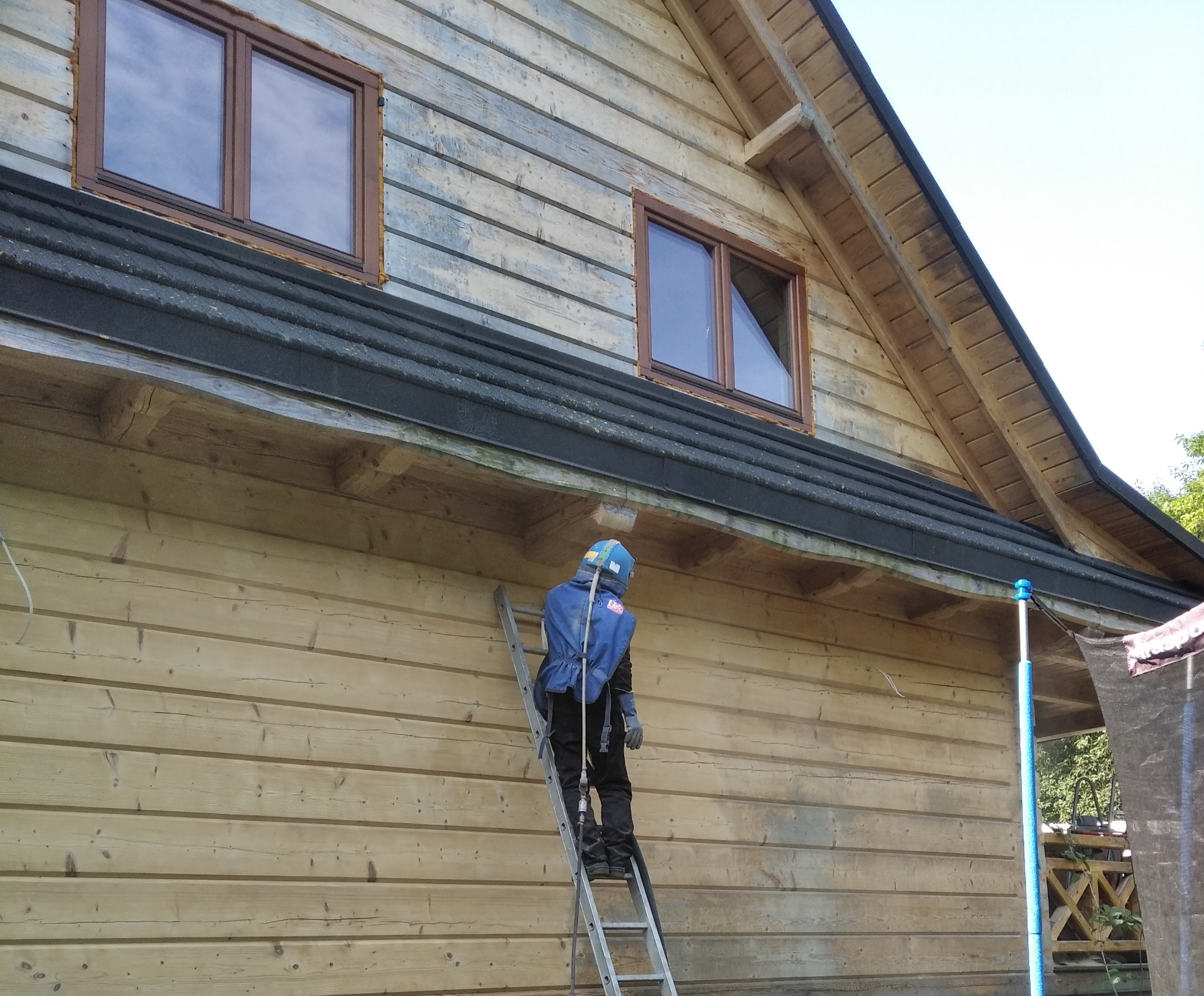 Pracownik w kombinezonie ochronnym na drabinie, przygotowujący drewnianą elewację domu do malowania, widoczne okna z brązowymi ramami i fragment dachu.