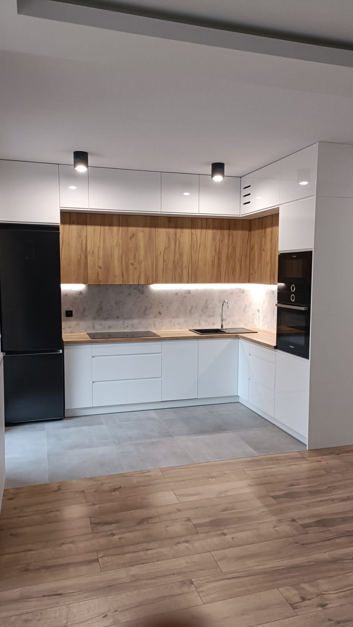 Nowoczesna kuchnia z białymi szafkami dolnymi i drewnopodobnymi górnymi, czarny sprzęt AGD, szara ściana nad blatem i podświetlenie LED, podłoga z płytek imitujących drewno.