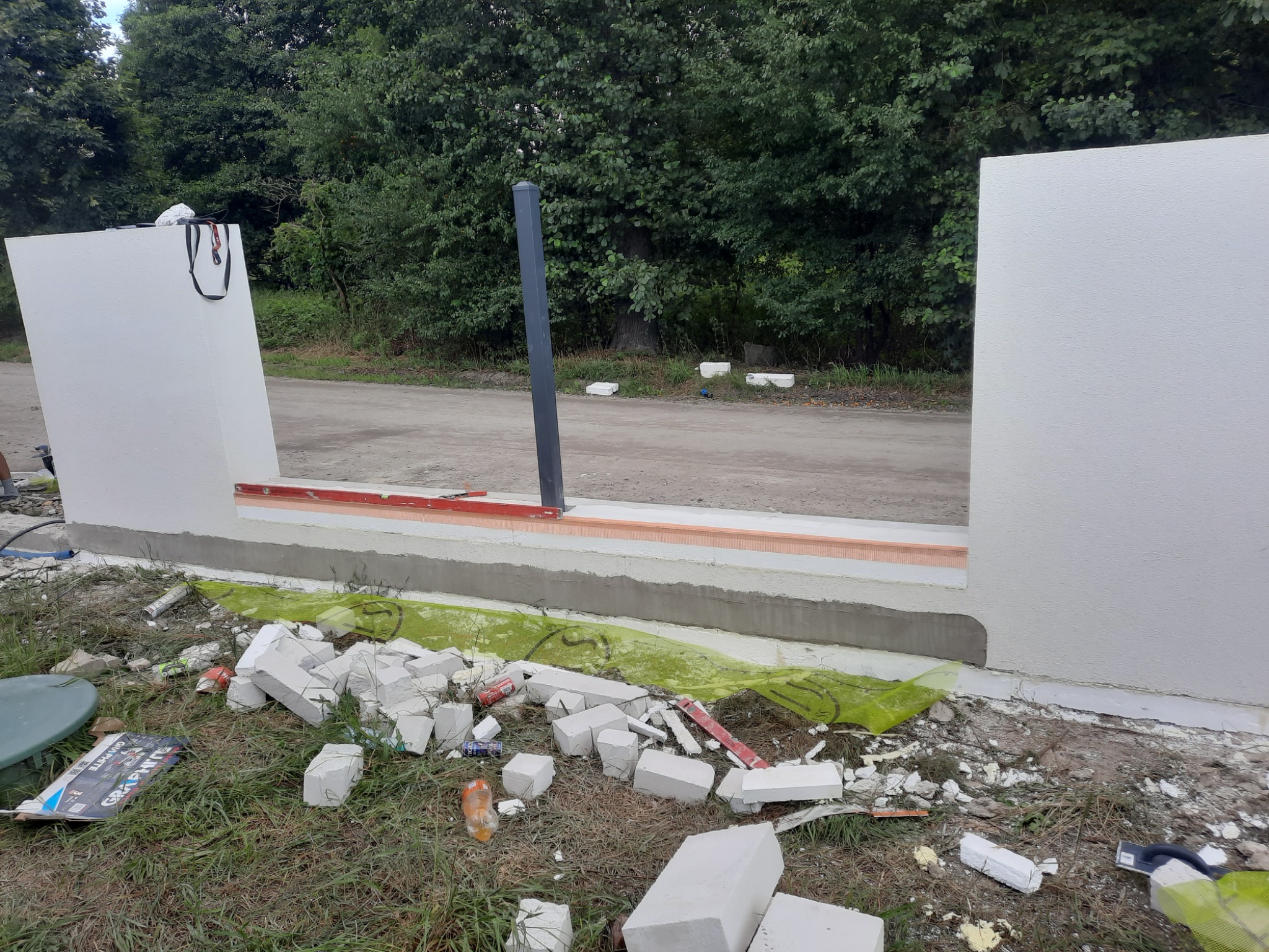Wznoszenie ściany z białych bloczków na fundamencie z wylewką, widoczne resztki materiałów budowlanych i narzędzia na trawie, w tle drzewa i droga.