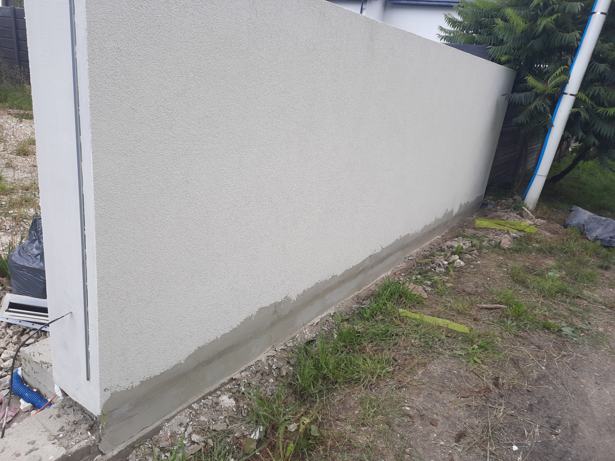 Szara ściana zewnętrzna z nałożoną strukturą, widoczna dolna partia z szarą warstwą wykończeniową, obok gruz i resztki materiałów budowlanych.