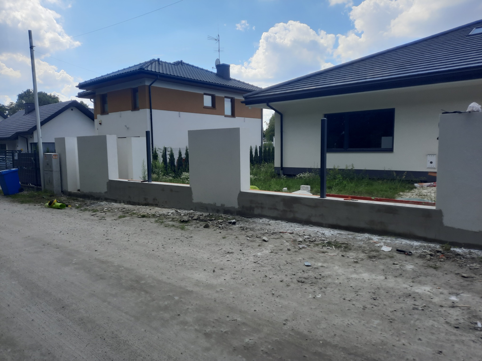 Budowa ogrodzenia z betonowych słupków i podmurówki na tle domów jednorodzinnych w słoneczny dzień.