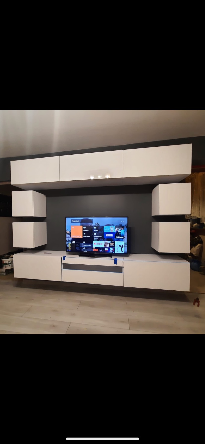 Biała, nowoczesna meblościanka z podświetleniem, zamontowana na szarej ścianie, z telewizorem z włączonym Netflixem, widoczne ślady taśmy malarskiej na szafce RTV.