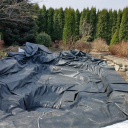 Bartosz Bolonek - Folia EPDM rozłożona w wykopie pod budowę oczka wodnego. W tle rząd tuj i inne rośliny ozdobne. Widoczne kamienie brzegowe i fragment ścieżki.