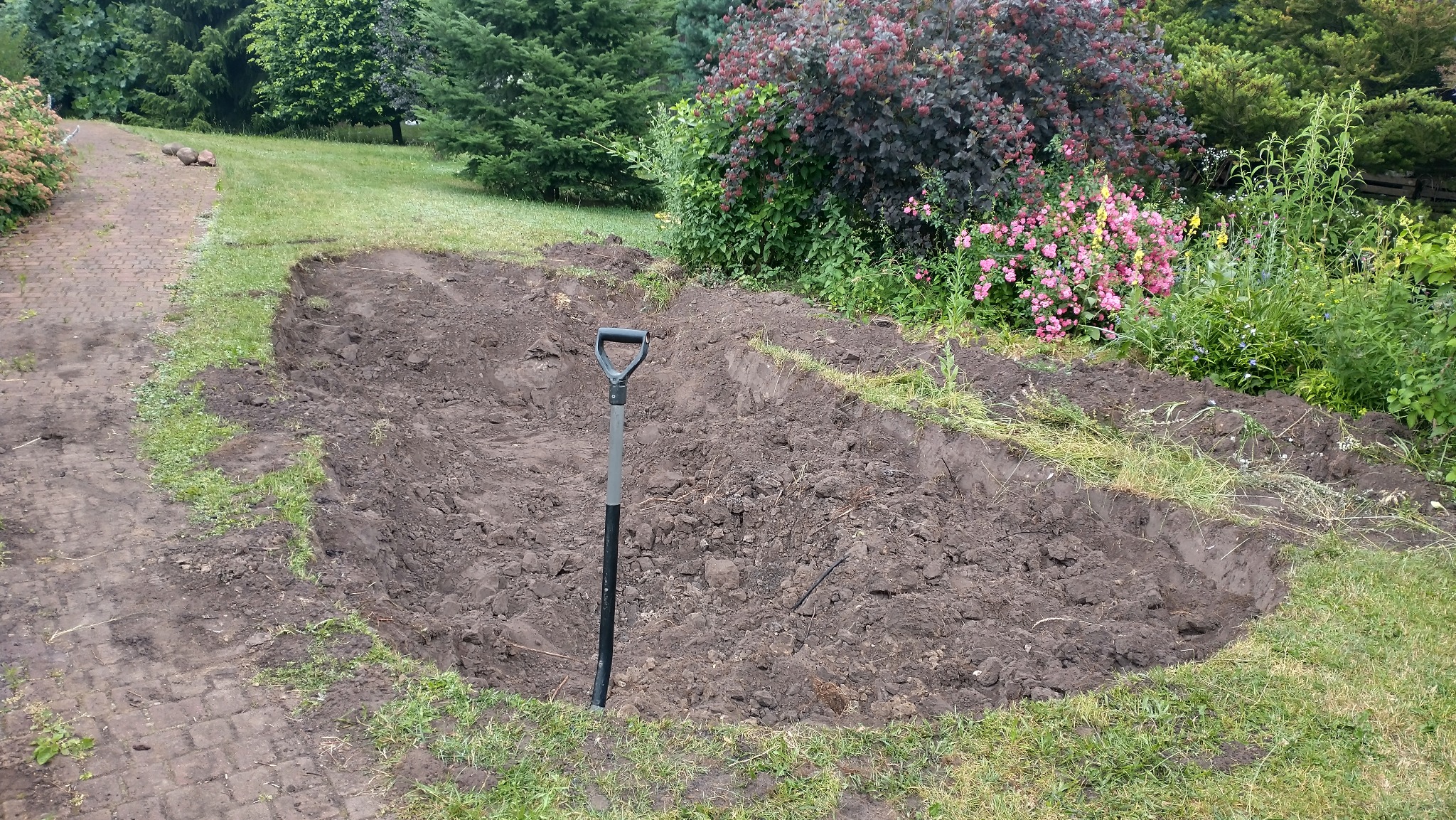 Wykopany dół w ziemi, prawdopodobnie pod oczko wodne, z wbita łopatą. W tle widoczna zieleń ogrodu i ścieżka z kostki brukowej. Etap prac ziemnych.