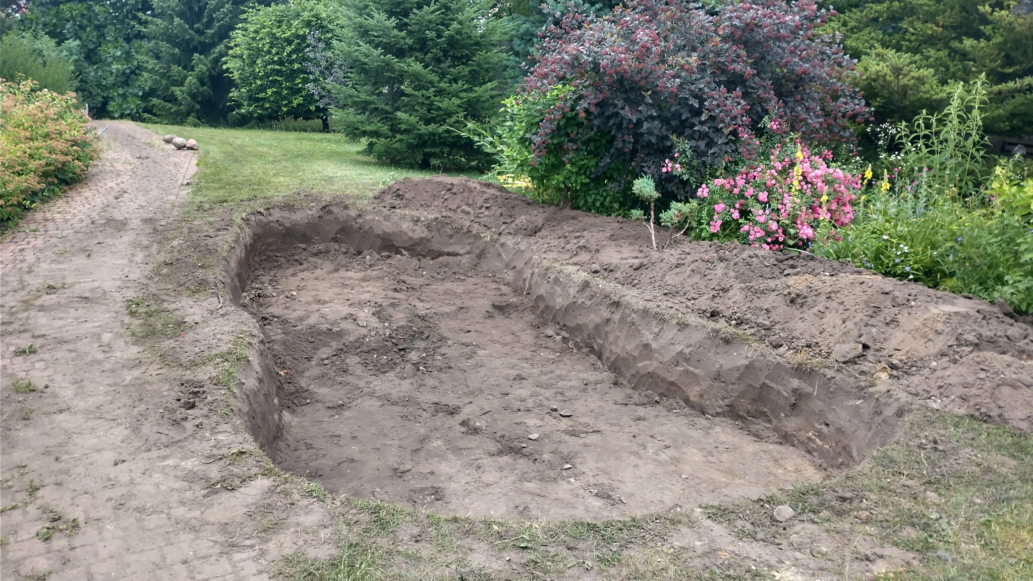 Wykonany wykop pod oczko wodne w ogrodzie, widoczna ziemia i roślinność w tle. Ścieżka z kamieni prowadzi wzdłuż trawnika.