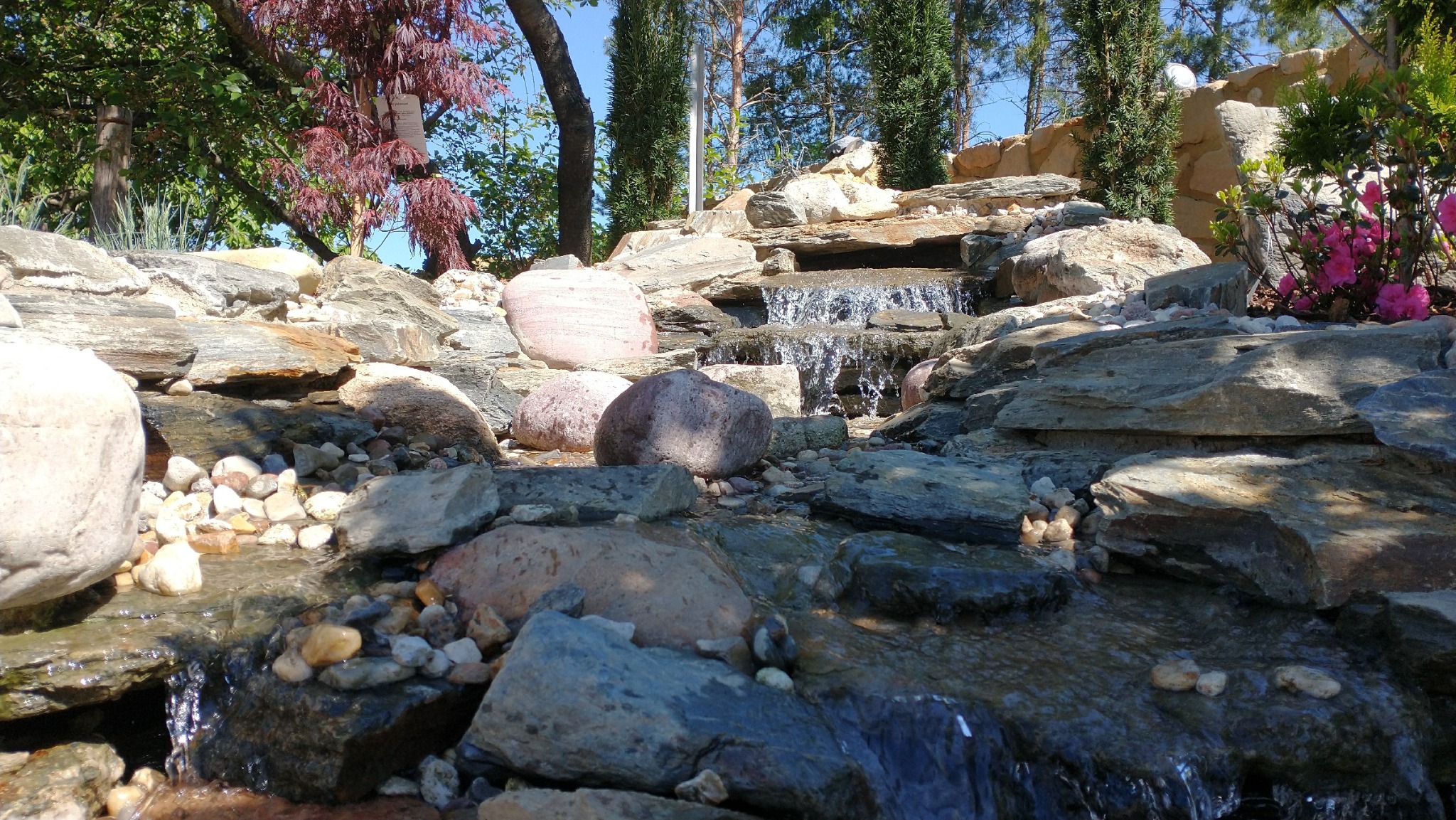 Kaskada wodna z kamieni w ogrodzie, z roślinami i drzewami w tle. Ujęcie z dołu, podkreślające naturalny charakter aranżacji.