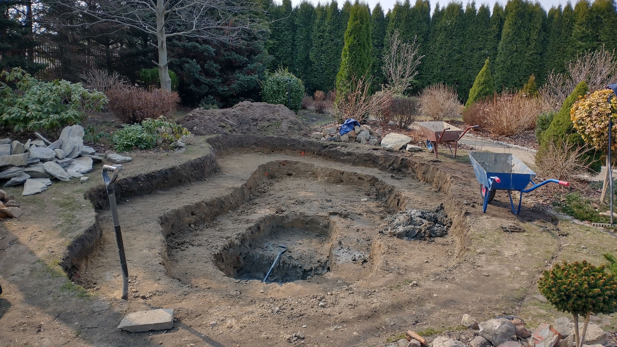 Wykopany dół pod oczko wodne w ogrodzie, widoczne narzędzia i taczki. Etap prac ziemnych, przygotowanie terenu pod budowę zbiornika wodnego.