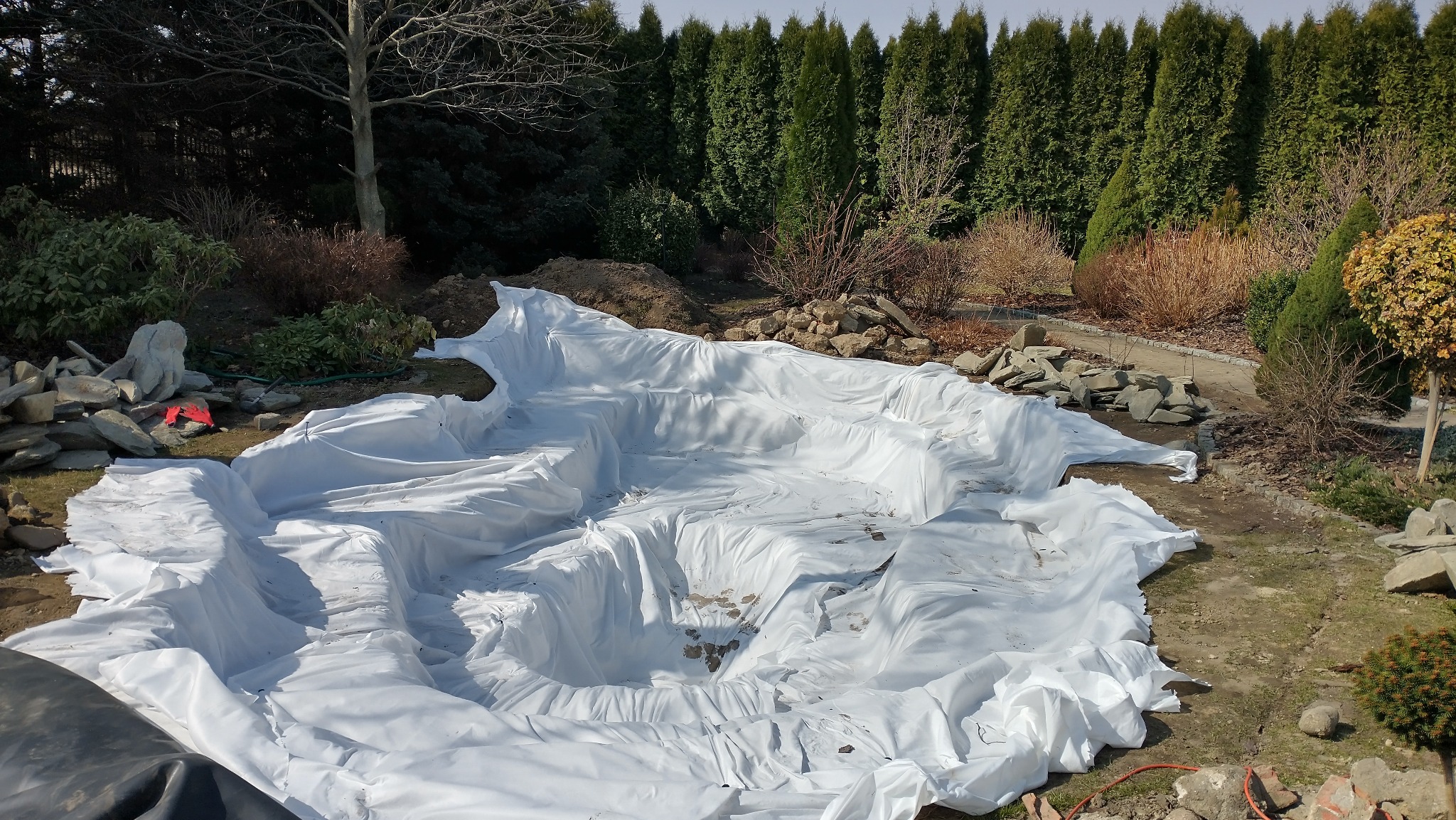 Etap budowy oczka wodnego w Pabianicach: biała folia ułożona w wykopie, kamienie i roślinność w tle. Widok z góry, słoneczny dzień.
