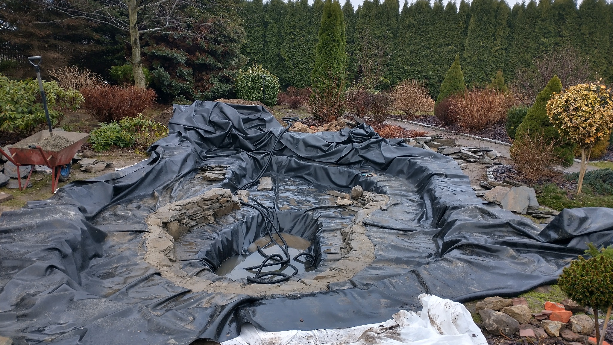Budowa oczka wodnego: wykop wyłożony czarną folią, częściowo wypełniony wodą, z kamieniami na brzegu. W tle zielone tuje i inne rośliny.