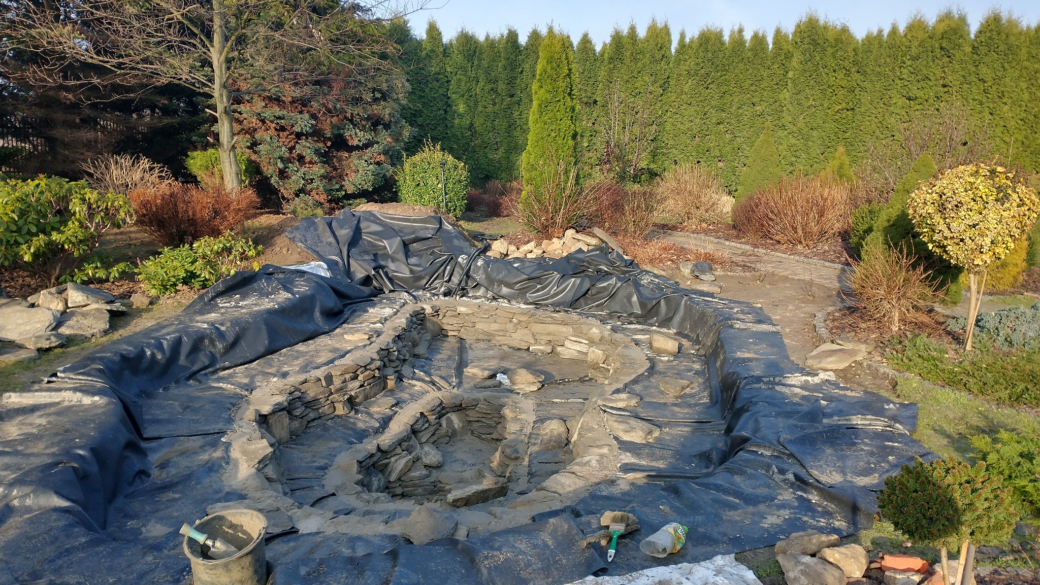 Budowa oczka wodnego w ogrodzie: wykop wyłożony czarną folią, kamienne obrzeża w trakcie tworzenia, narzędzia pracy obok. W tle tuje i krzewy.