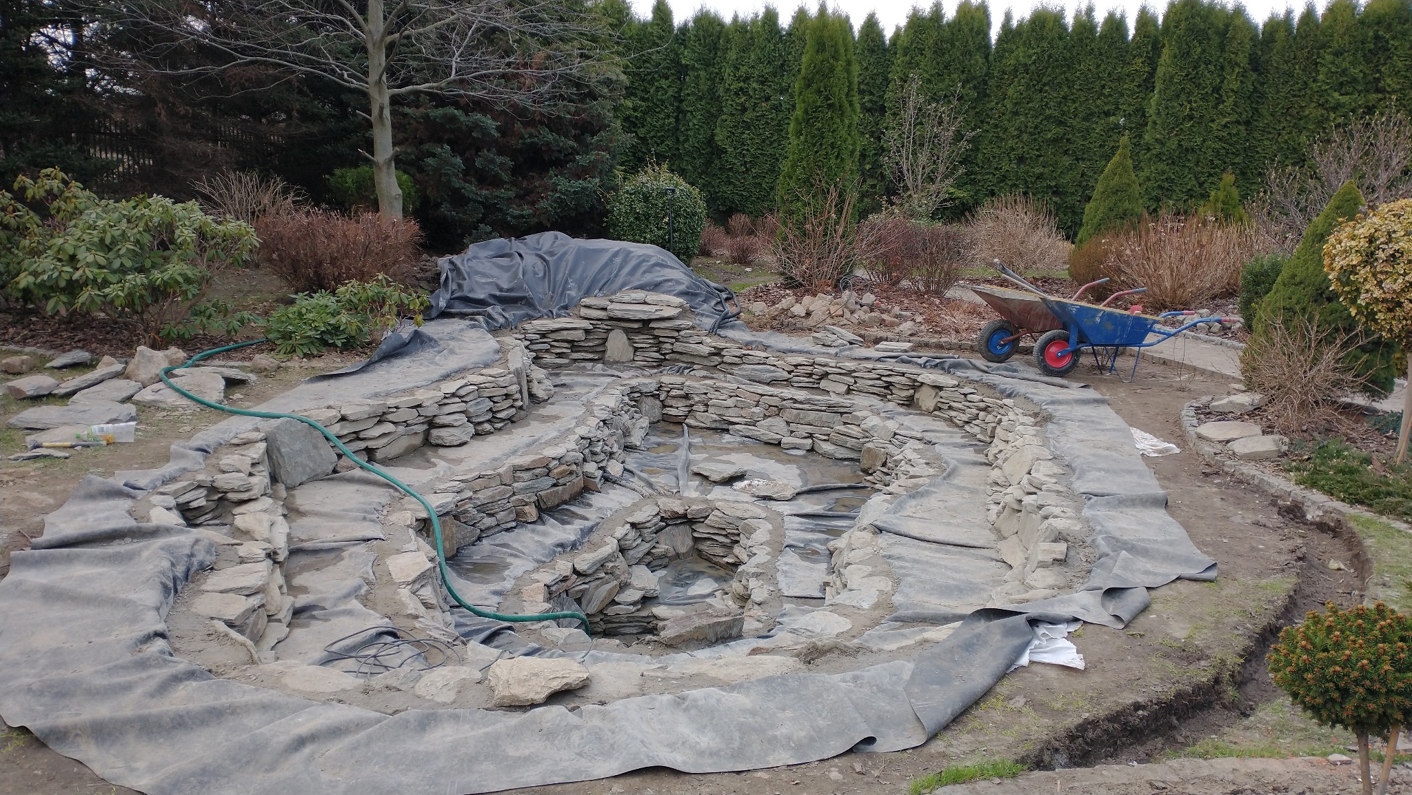 Budowa kaskadowego oczka wodnego z kamienia w ogrodzie. Widoczne etapy prac, folia ochronna, kamienie i dwie taczki w tle. Ujęcie z góry, w dzień.