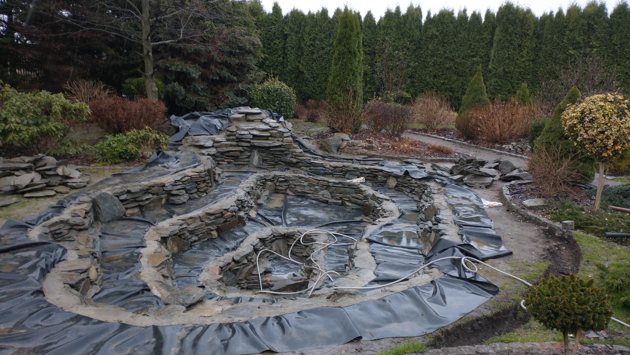 Budowa kaskadowego oczka wodnego z kamienia, wyłożonego czarną folią, otoczonego zielenią. Widoczne etapy prac ziemnych i układania kamieni.