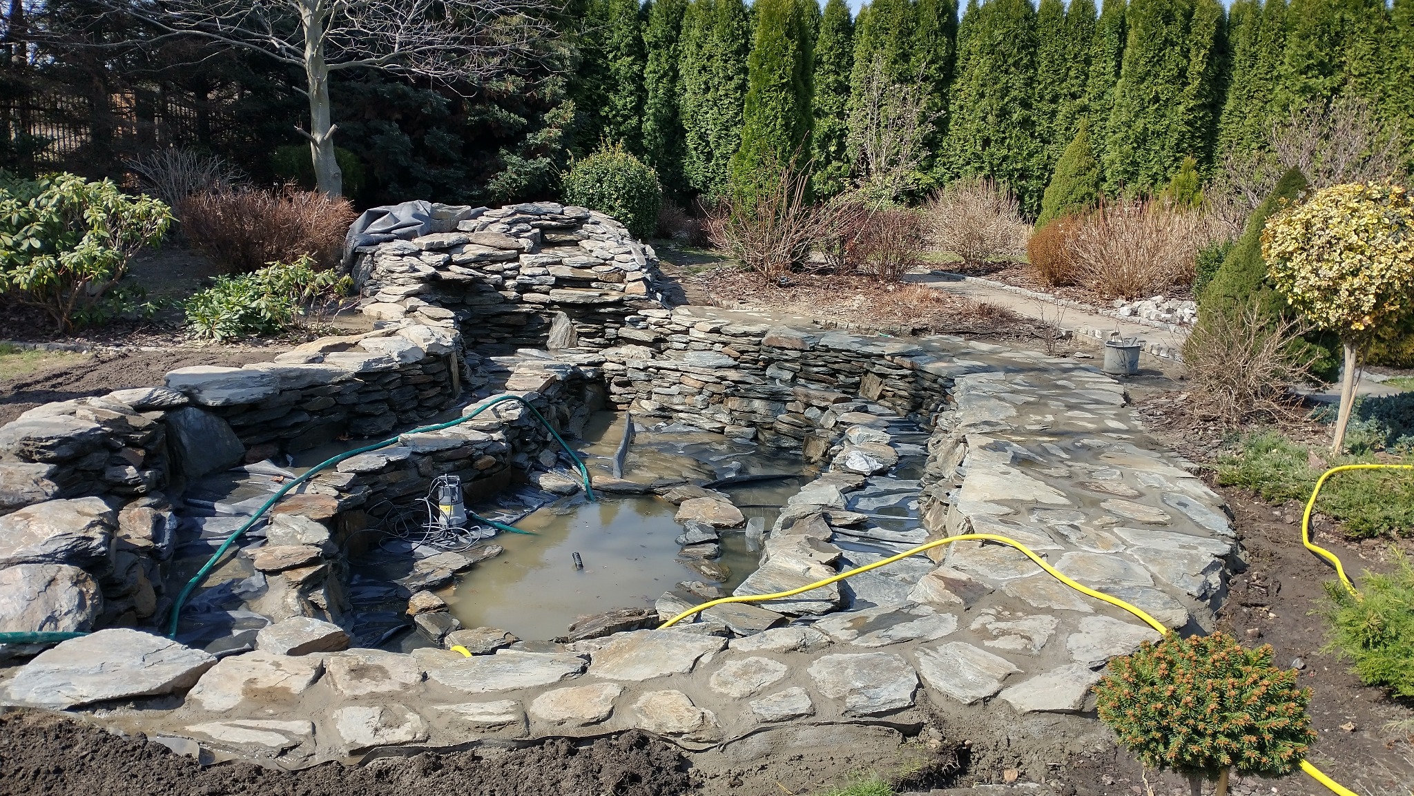 Kamienny zbiornik wodny w ogrodzie, z kaskadą i roślinnością w tle. Woda lekko zabrudzona, widoczne węże i pompa. Etap budowy lub czyszczenia.