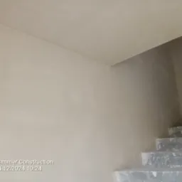 Świeżo otynkowana, gładka ściana w odcieniach beżu z widocznymi surowymi, betonowymi schodami w tle, ujęcie z dołu.