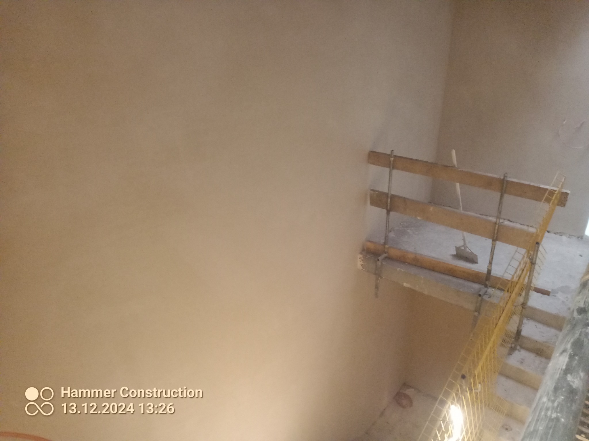 Świeżo nałożone tynki na ścianie klatki schodowej, widoczne rusztowanie z żółtą siatką zabezpieczającą.