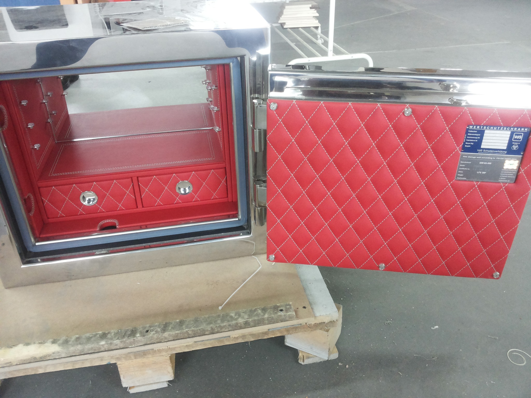 Otwarty sejf z chromowanymi elementami i czerwonym, tapicerowanym wnętrzem w romby, widoczny na drewnianej palecie.