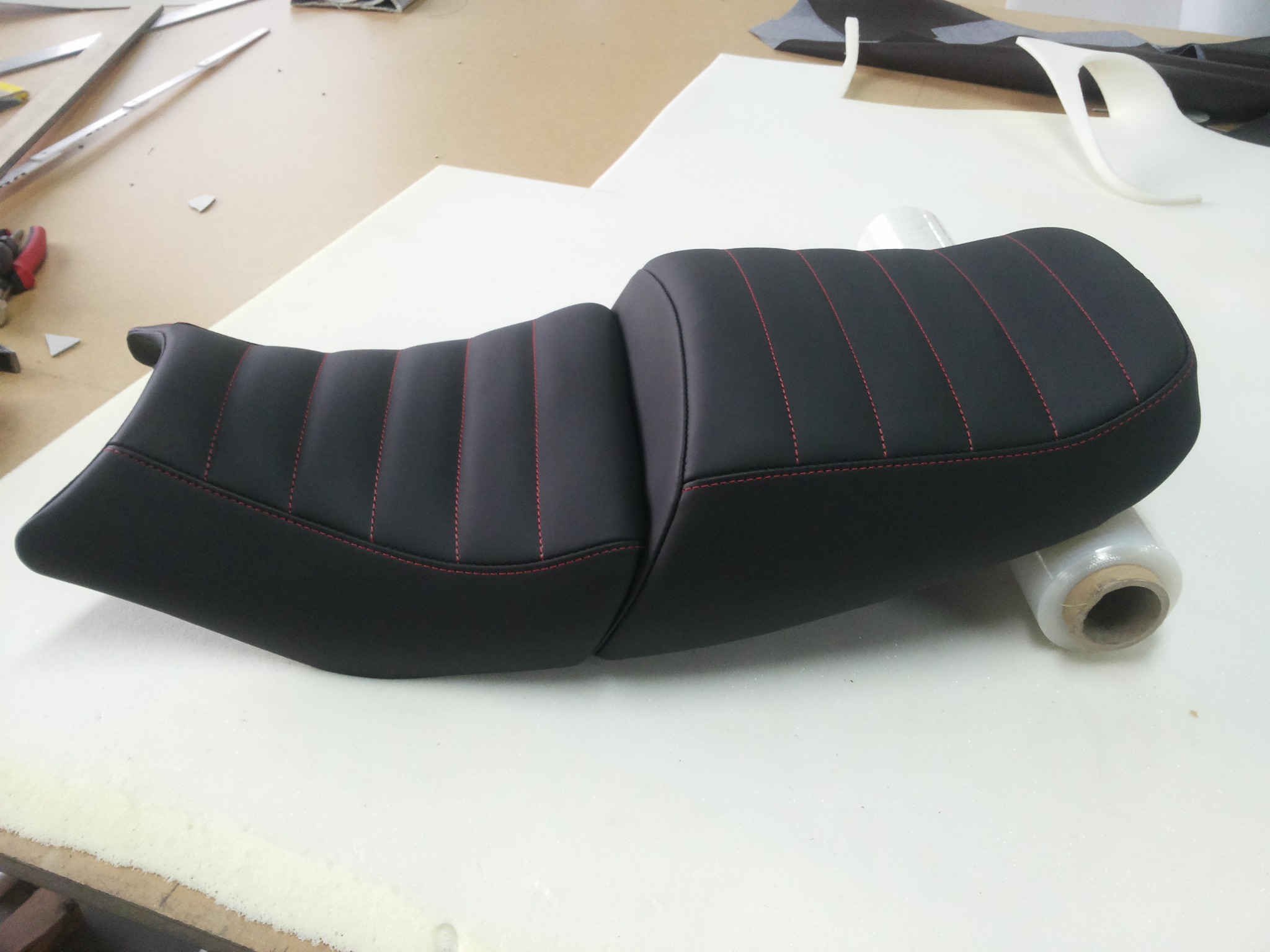 Czarne, skórzane siedzenie motocyklowe z czerwonymi przeszyciami na białym podłożu, częściowo owinięte folią stretch.