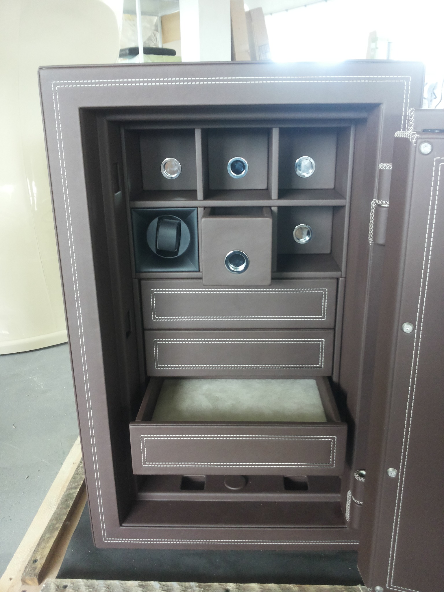 Otwarty sejf wykończony brązową skórą z białymi przeszyciami, widoczne półki, szuflady i nawijarka do zegarków.