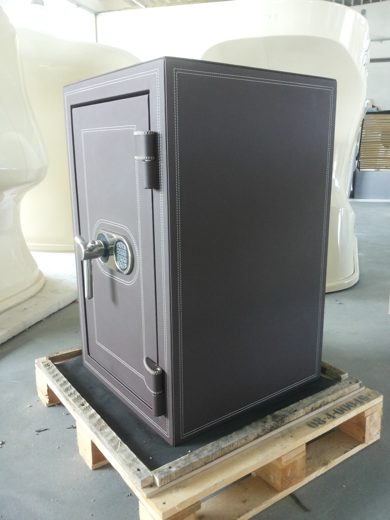 Brązowy sejf obity skórą z elektronicznym zamkiem, ozdobne przeszycia na krawędziach, umieszczony na drewnianej palecie.