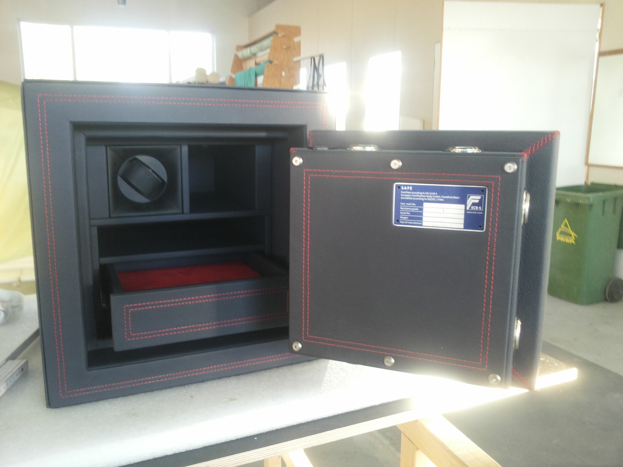 Otwarty sejf obity czarną skórą z czerwonymi przeszyciami, widoczna wewnętrzna półka z czerwonym dnem i mechanizm do nakręcania zegarków.